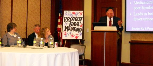 Leighton Ku presents next to a sign that reads "Protect Kids' Medicaid." 